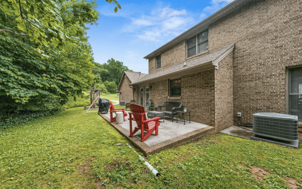 Backyard patio with seating and greenery beside a brick house.