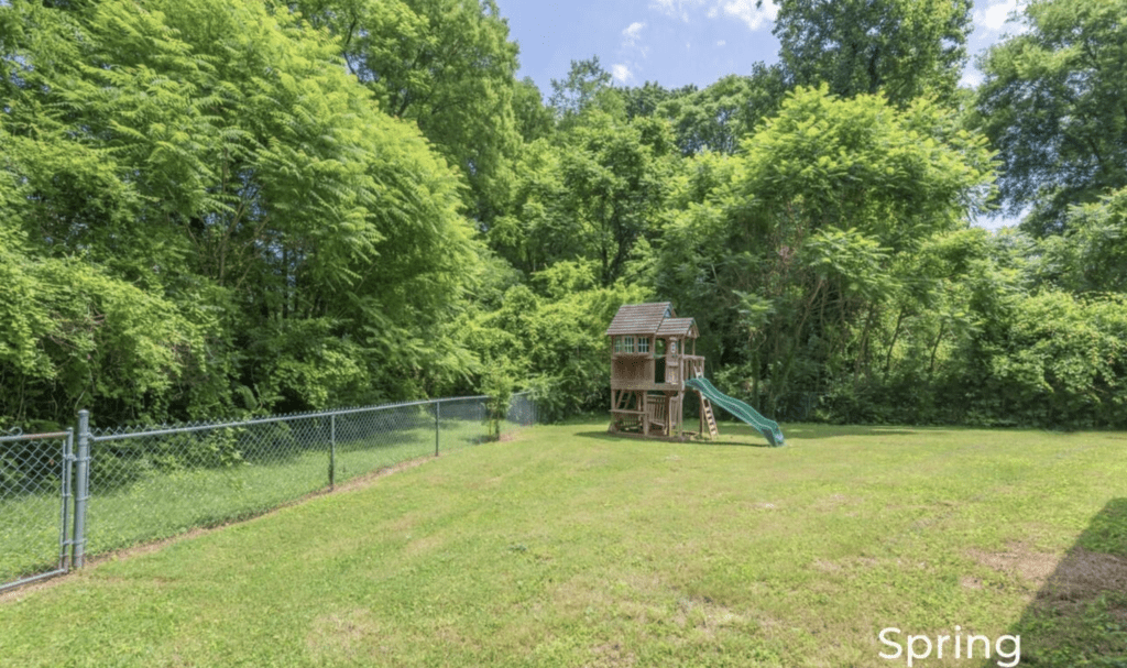 A wooden playset with a slide in a grassy backyard surrounded by trees.
