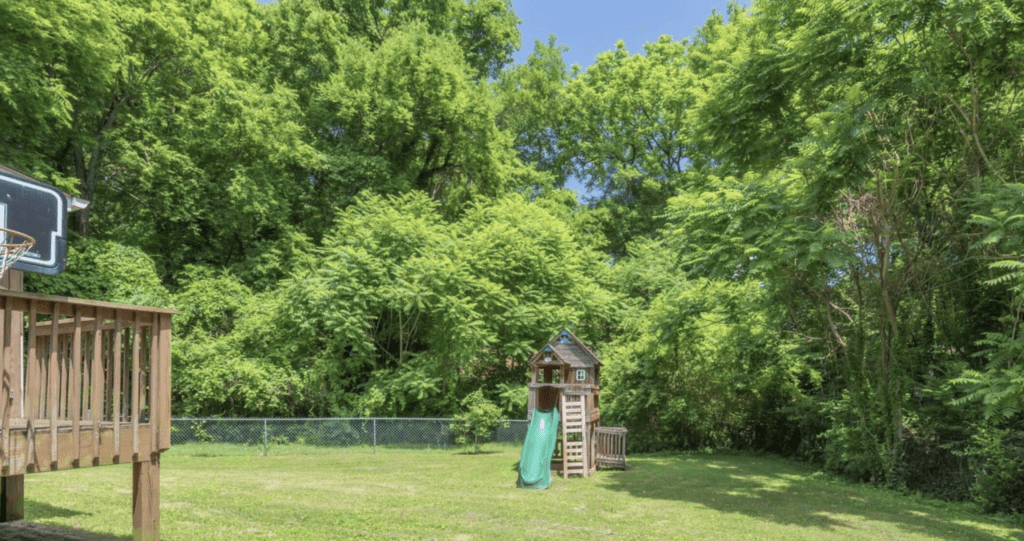 A wooden playground set with a slide in a grassy yard surrounded by trees.